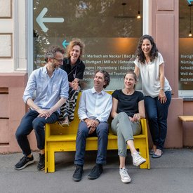 Das Team der Geschäftsstelle der KulturRegion Stuttgart sitzt auf einer gelben Bank vor der Geschäftsstelle.