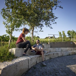 Zwei Personen, möglicherweise ein Paar - sitzen auf halbrund angeordneten Steinen und unterhaten sich. Im Hintergrund ist ein eg durch die Weinberge zu sehen.