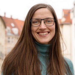 Portrait von Tina Pantisano. Sie hat langes, glattes, braunes Haar, trägt eine Brille und einen türkisfarbenen Rollkragenpulli. Tina Pantisano lächelt freundlich in die Kamera.