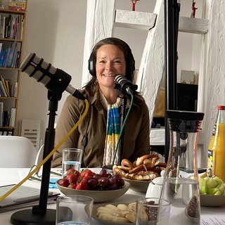 Gästin Dr. Cornelie Kunkat (Deutscher Kulturrat) lächelnd bei der Podcast-Aufnahme. Sie trägt Kopfhörer und sitzt vor einem Tischmikrofon. Vor ihr auf dem Tisch befindet sich noch mehr Podcast-Technik und verschiedene Snacks und Getränke.
