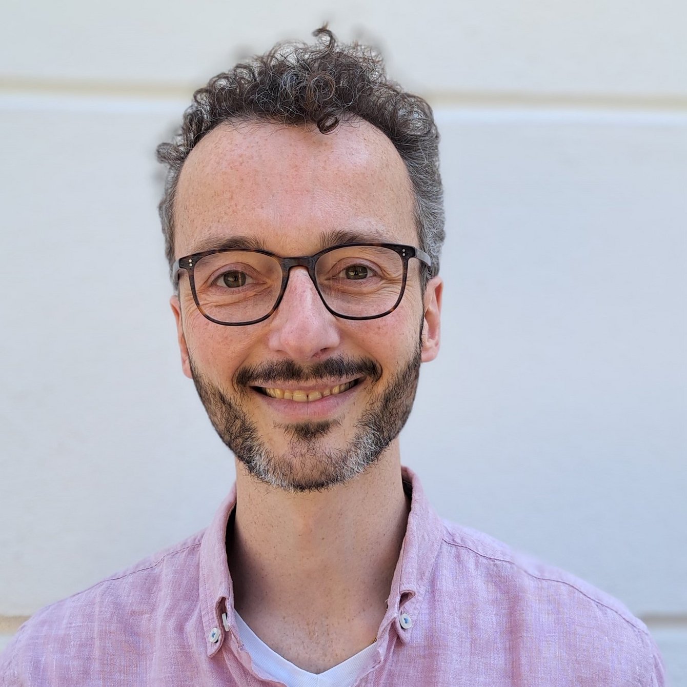 Portraitfoto von David Studniberg. David Studniberg hat dunkle Locken, einen kurzen Bart und trägt eine schwarze Brille. Bekleidet ist er mit einem rosa Hemd. Er lächelt freundlich und offen.