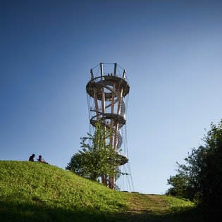 Foto: Blick von unten über eine grüne Wiese hinweg auf den Turm. Auf der Wiese sitzen zwei Personen. Der Himmel ist blau.