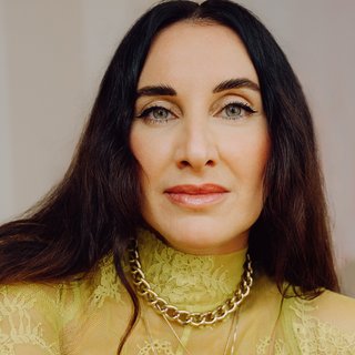 Portrait von Mirna Funk: Mirna hat lange schwarze Haare, sie trägt eine gelbe Spitzenbluse und verschieden Halsketten.