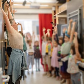 Eine Gruppe Kinder strecken ihre Arme offensichtlich nach Anleitung einer erwachsenen Person am linken Bildrand Richtung Decke.