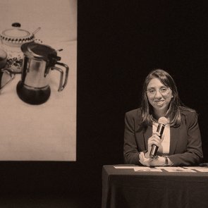 Karina Pino Gallardo sitzt an einem Tisch und hält ein Mikrofon in der Hand. Hinter ihr ist ein Bild mit einem Kaffeekännchen, einer Kaffeetasse, einem Löffel und einer Zuckerdose zu sehen.