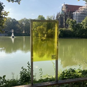 Bild vom Festival Floß auf dem Feuersee in Stuttgart. Fotografiert vom Ufer des Sees durch eine gelbliche Scheibe hindurch. Im Hintergrund die Johanneskirche.