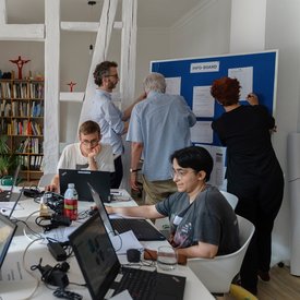 Zwei Personen sitzen mit ihren Laptops an einem Tisch und scheinen zu arbeiten. Im Hintergrund stehen drei Personen an einem Flipchart