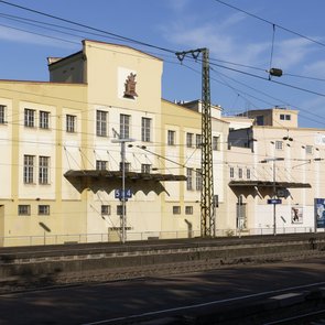 Blick auf die Aussenansicht des Franck-Areals über Gleise hinweg. Das alte Firmengebäude wird von der Sonne angestrahlt, darüber blauer Himmel.