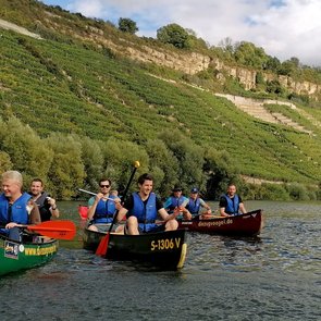 Bild von mehrere mit Personen besetzte Kanus auf dem Neckar. Im Hintergrund ein steiles Ufer mit Weinbergen und Felsen.