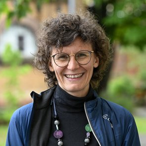 Portrait von Lucia Leidenfrost. Lucia Leidenfrost hat eher kurz geschnittene Locken und trägt eine Brille. Bekleidet ist sie mit einem schwarzen Rollkragenpulli, einer blauen Jacke und einer auffälligen, langen Perlenkette. Sie lächelt offen in die Kamera.