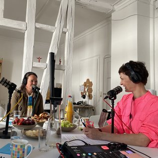 Gästin Dr. Cornelie Kunkat (Deutscher Kulturrat) und Moderatorin Kimsy von Reischach bei der Podcast-Aufnahme. Sie tragen beide Kopfhörer und haben je ein Tischmikrofon vor sich. Sie scheinen miteinander zu sprechen und lächelnd einander zu.