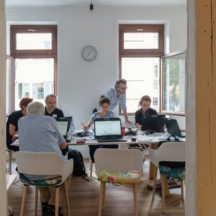 Eine Gruppe Menschen sitzt an einem Tisch und arbeitet an Laptops. Eine Person, David Studniberg, steht hinter einer Person und scheint dieser etwas zu erklären