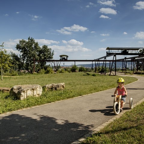 Bild: ein kleines Kind fährt mit einem Dreirad auf einem Weg durch einen Park. Ein Mann, vermutlich der Vater, fährt mit dem Roller hinter dem Kind. Im Hintergrund sind alte Bergbauanlagen zu sehen und es eröffnet sich ein Blick in ein Tal.