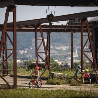 Kinder fahren Fahrrad zwischen stillgelegten Bergbauanlagen im Travertinpark. Ein Mann, vermutlich der Vater, sitzt auf einer Bank und schaut den Kindern zu. Im Hintergrund öffnet sich der Blick auf ein Tal.